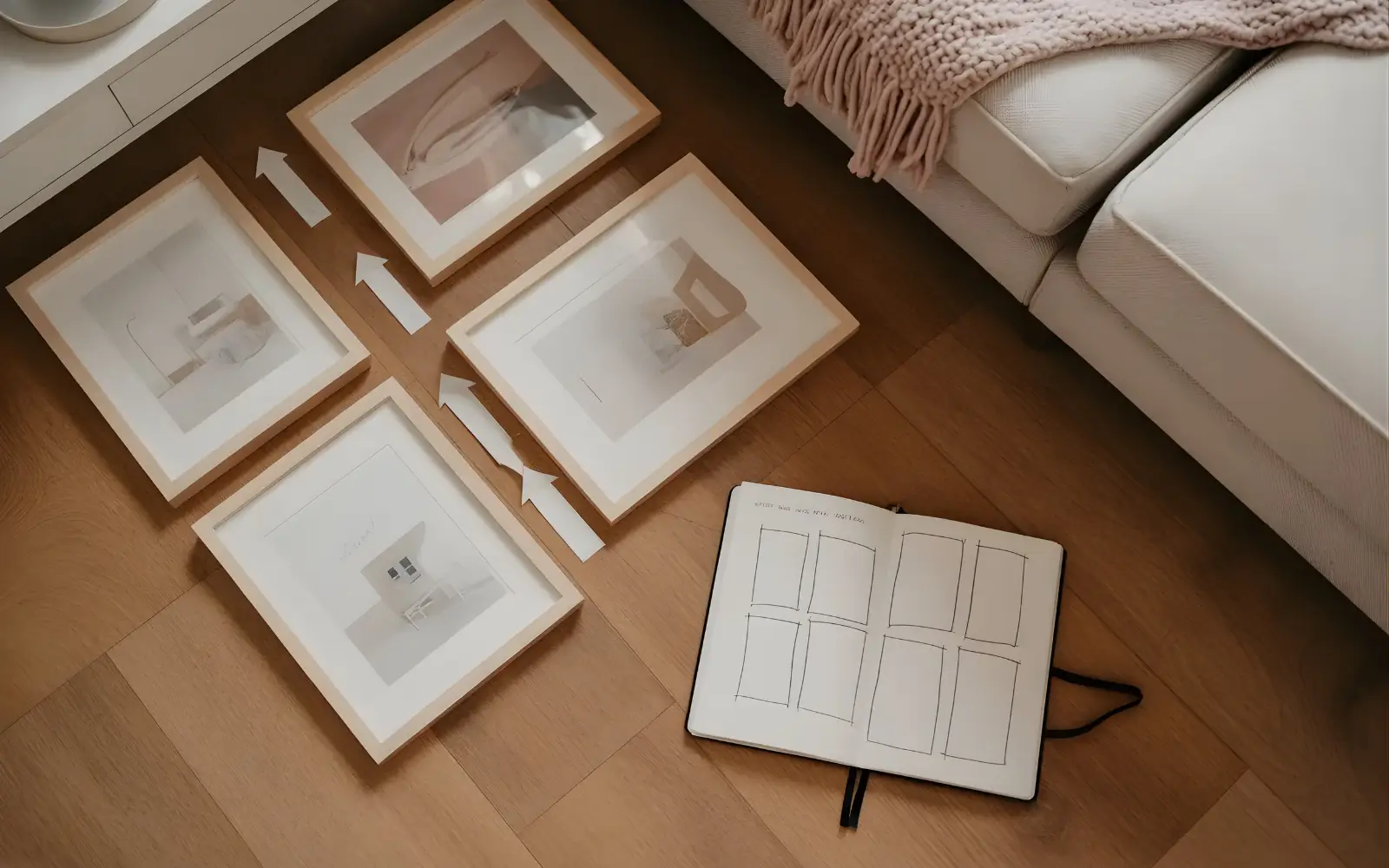 Overhead view of framed art laid out on a wooden floor in front of a sofa, with even gaps and a sketchbook showing the planned gallery wall layout.