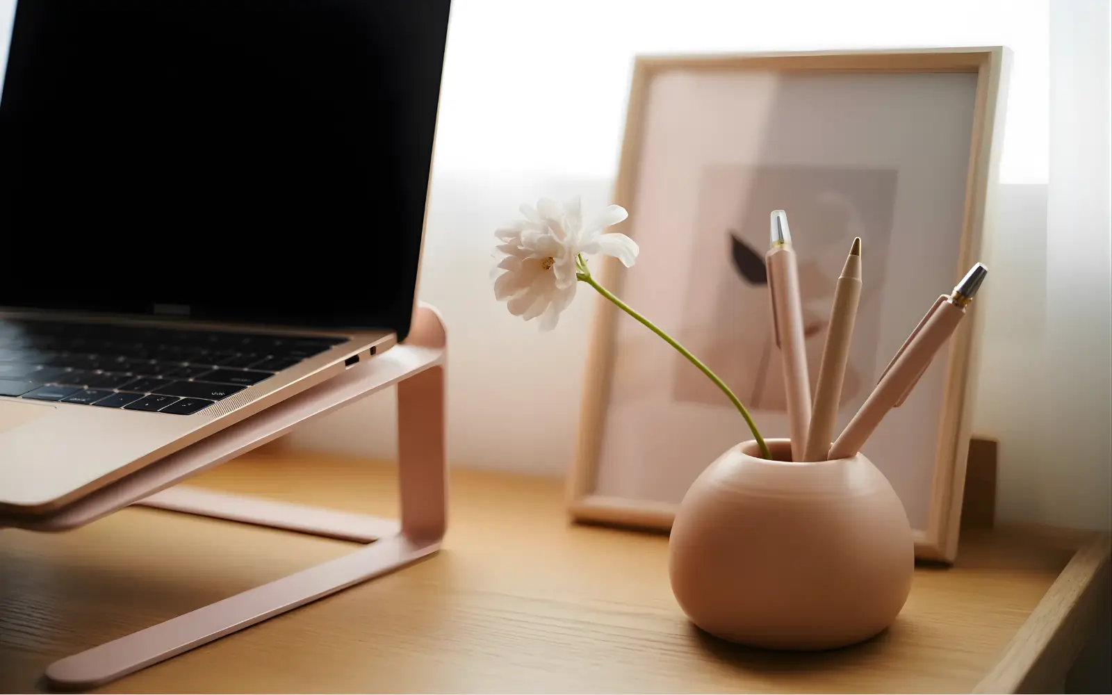 Blush and camel feminine desk setup with a laptop on a matching stand, ceramic pen cup, small bud vase and framed art in the background, styled as a curated desktop.